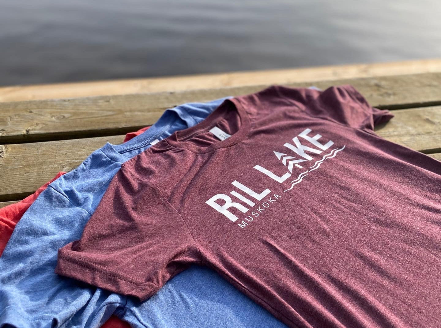 3cotton t-shirts displayed on a dock. T-shirt colours are maroon with white HTV log that reads RIL LAKE MUSKOKA, light blue heather and red heather.
