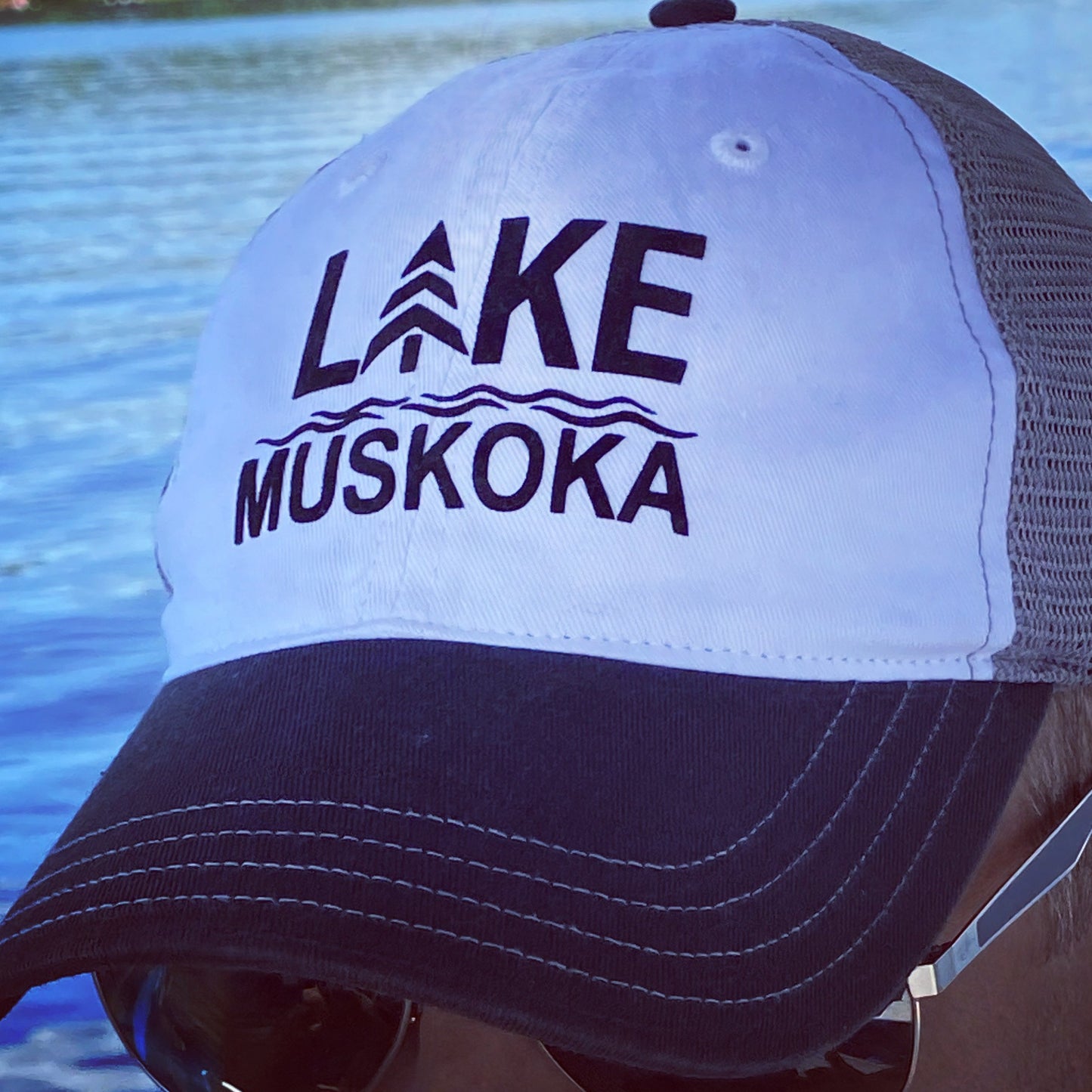 Male model wearing unisex cap with black/white cotton front and charcoal mesh back. HTV logo in black reads LAKE MUSKOKA.