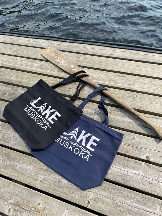 Custom LAKE logo large zippered, canvas tote bag. Shown in navy blue and black with white HTV logo that says AT THE LAKE MUSKOKA. Displayed by the water on  a dock with a canoe paddle.