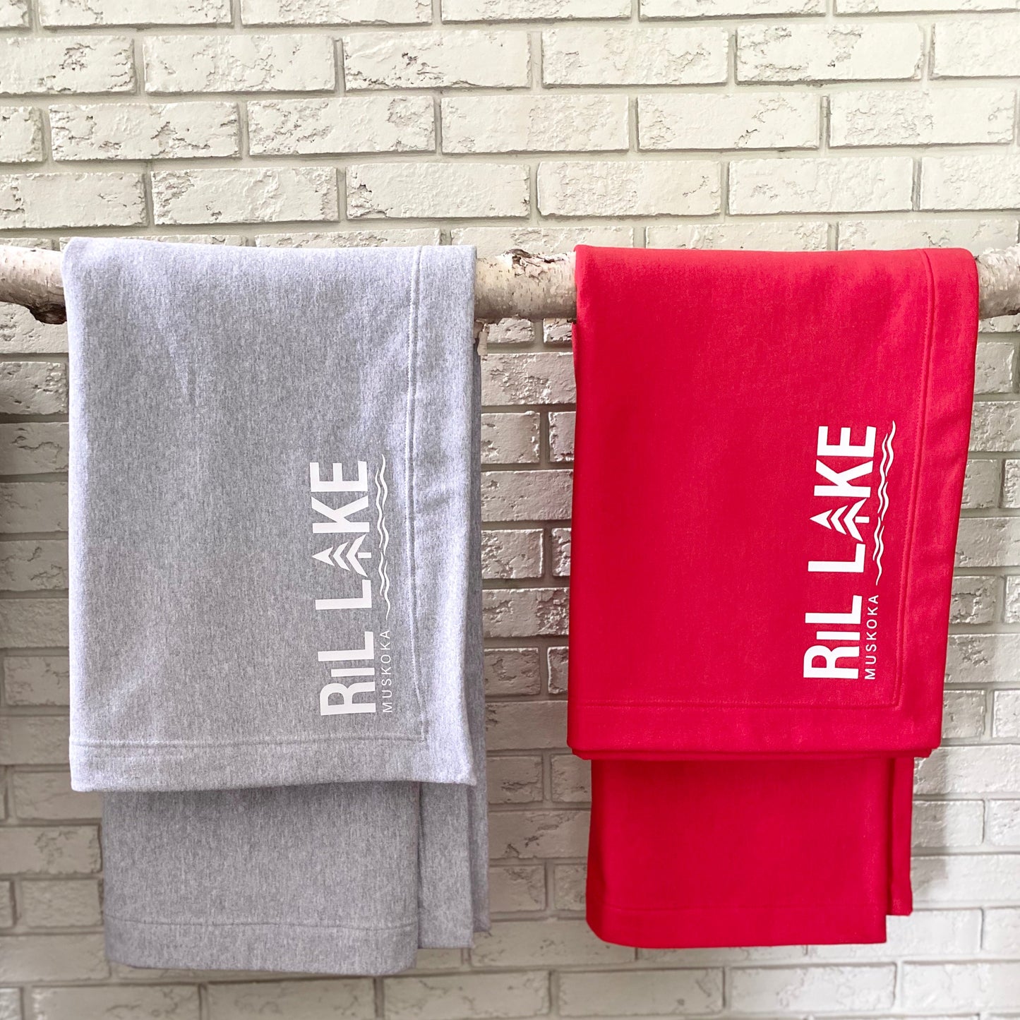 Sport grey heather fleece throw with white HTV  Ril Lake logo and Red  fleece throw blanket with white HTV Ril Lake logo. Both are hanging folded over a horizontal birch poled.