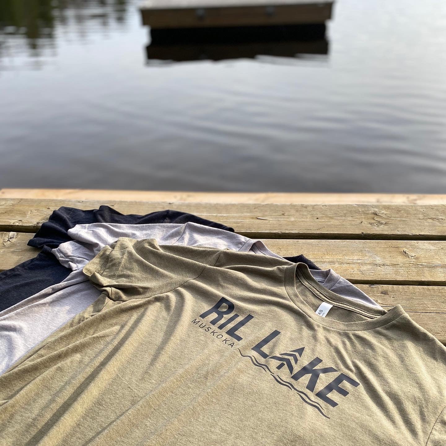 3 cotton t-shirts displayed  laying flat on a dock. Colours are olive with black logo that reads RIL LAKE MUSKOKA, sport grey and black heather.